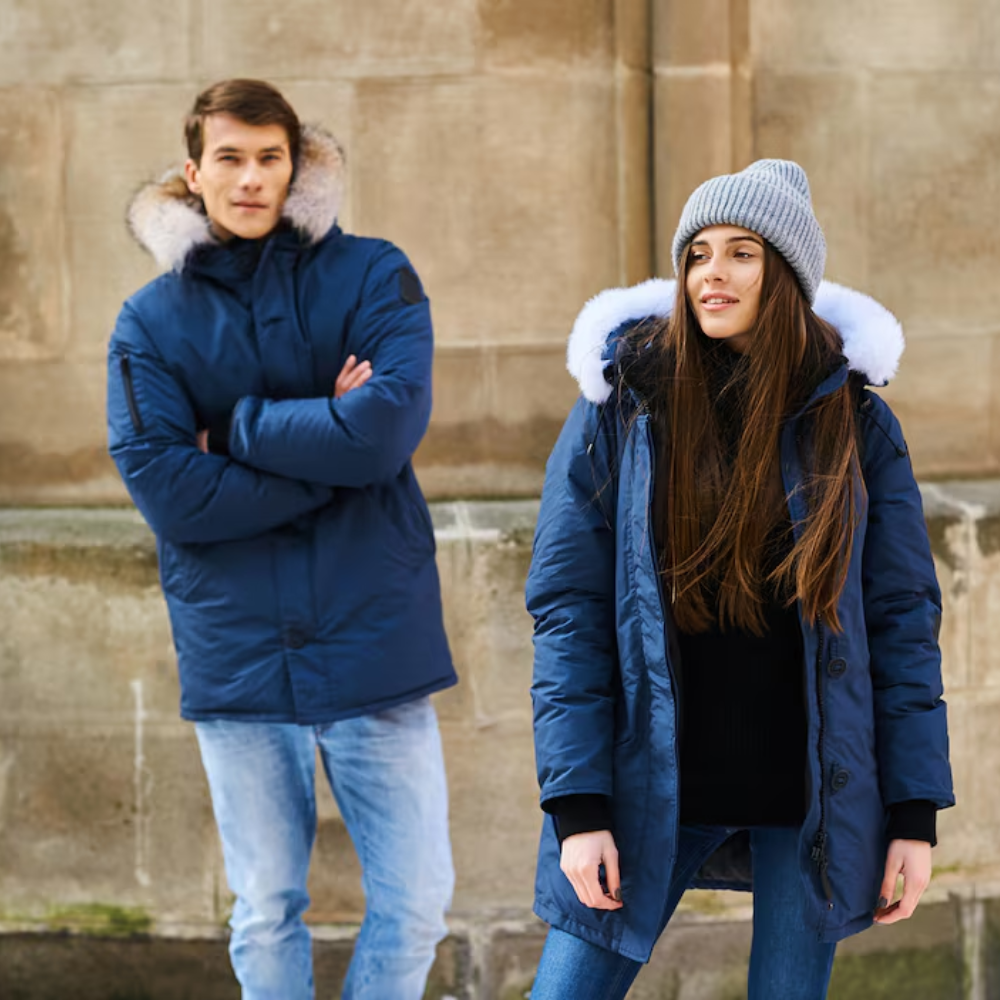 Two people wearing blue winter coats with fur-lined hoods standing against a stone wall.

